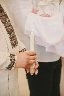 Nakışlı gömlekli bir adam yanan bir mum tutuyor, bir bebeğin vaftiz töreninde ışığı ve inancı simgeliyor.