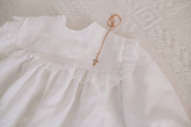 Beyaz bebek vaftiz elbisesi, ince dantel detaylar ve dini bir vaftiz törenini sembolize eden küçük bir altın haç kolyesi.