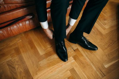 Man getting ready for a formal event, tying up his black leather shoes on a parquet floor with a leather sofa