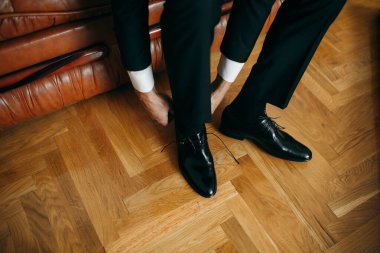 Elegant man in black suit pants and white shirt cuffs tying shoelaces of his formal shoes on a wooden floor