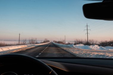 Sürücünün kar ve elektrik direkleriyle çevrili uzun, temiz bir asfalt yola bakış açısı sakin bir kış gökyüzü altında