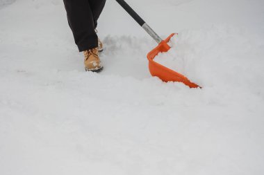 Kışlık botlar giyen, açık havada çalışan, yoğun bir kar yağışı sonrası turuncu bir kürekle derin karları temizleyen kişi.