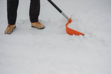 Kahverengi çizme ve siyah pantolon giyen dışarıda turuncu plastik bir kürekle kar küreyen kişi.