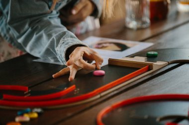 Çocuğun eli ahşap bir masada pembe bir oyun taşıyla meşgul, kırmızı raylarla eğlenceli bir masa oyunu oynuyor.