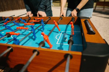 Eller langırt masasının çubuklarıyla meşgul, oyuncuları canlı bir açık hava oyunu sırasında kontrol ediyor, rekabeti ve eğlenceyi sembolize ediyor