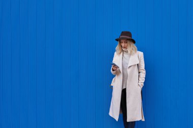 Şapkalı çekici bir kadın mavi arka planda cep telefonuyla mesaj yolluyor.