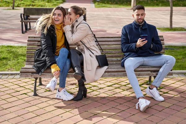 2人の美しいティーンエイジャーが秘密を共有し 噂話をしながら男の子は彼の携帯電話を見て 彼らは公園のベンチに座っている — ストック写真
