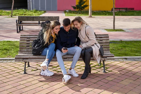 屋外に座って携帯電話を見ている3人の若い友人 屋外の公園のベンチに座って スマートフォンでビデオを見ている人々のグループ 友情と技術の概念 — ストック写真