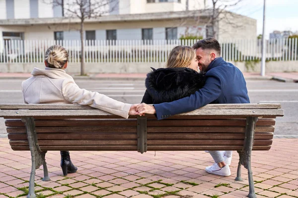 Love triangle, a girl kiss at her boyfriend and he shakes hands with another girl, they are sitting on a bench. Concept of Infidelity