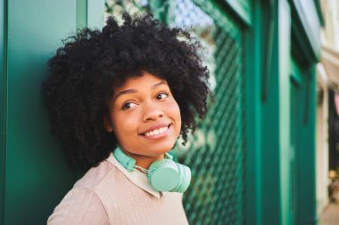Yeşil duvara yaslanmış kulaklıklı, gülümseyen afro saçlı kadın. Mutlu Latin kadın portresi 
