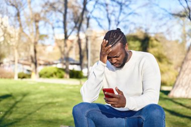 Parktaki bankta oturan Afro-Amerikalı adam cep telefonuna bakıyor. Endişeli genç adam. Teknoloji kavramı