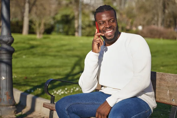 Homem negro feliz falando no telefone inteligente sentado em um parque em um dia ensolarado. — Fotografia de Stock