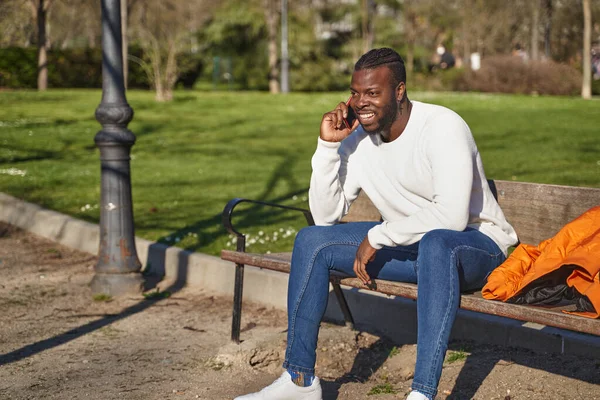 Pria kulit hitam yang bahagia berbicara di telepon pintar duduk di taman di hari yang cerah. — Stok Foto