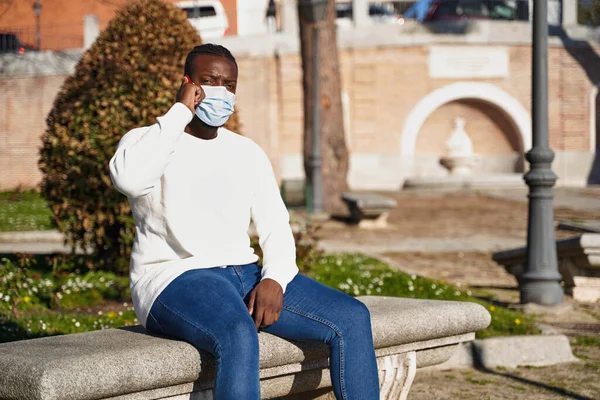 Un giovane afroamericano seduto su una panchina a parlare al telefono. Un uomo latino con la maschera anti-contagio. Concetto di nuova normalità. — Foto Stock