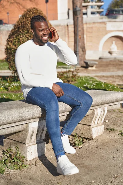 Homem negro feliz falando no telefone inteligente sentado em um parque em um dia ensolarado. — Fotografia de Stock