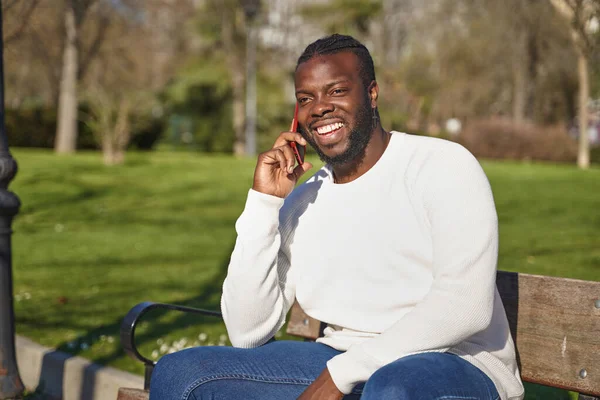 Güneşli bir günde parkta oturmuş akıllı telefondan konuşan mutlu siyahi adam.. 