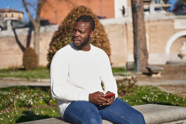 Pemuda Afrika Amerika duduk di bangku taman mengirimkan pesan. Pria latin dengan topeng anti-penularan. Konsep dari normal baru. — Stok Foto