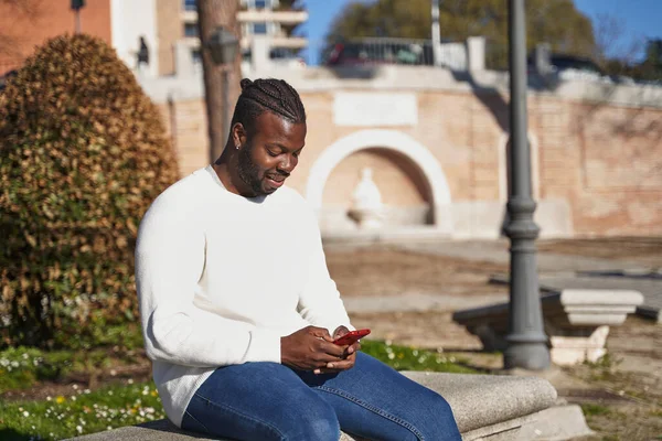 Jovem afro-americano sentado num banco do parque a enviar uma mensagem. Homem latino com máscara anti-contágio. Conceito de novo normal. — Fotografia de Stock