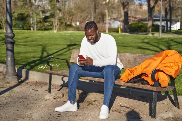 Pemuda Afrika Amerika duduk di bangku taman mengirimkan pesan. Konsep dari normal baru. — Stok Foto