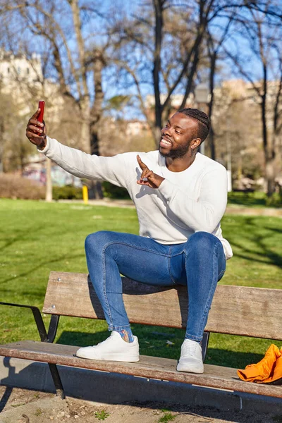 Jovem afro-americano sentado em um banco de estacionamento, tirando um auto-retrato com um smartphone. Homem negro feliz. Nova normalidade — Fotografia de Stock
