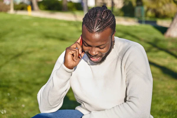 Homem negro feliz falando no telefone inteligente sentado em um parque em um dia ensolarado. — Fotografia de Stock