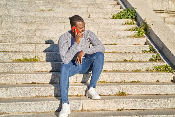 Pria kulit hitam yang senang bicara di telepon pintar duduk di tangga. Pemuda dengan celana jeans dan sweater abu-abu. Konsep komunikasi. — Stok Foto