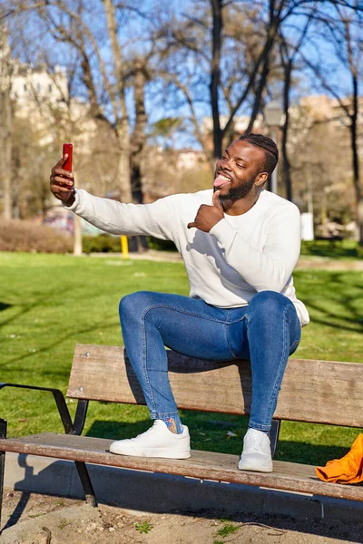 Un tânăr afro-american stând pe o bancă de parc, luând un autoportret cu un smartphone. Un negru fericit. Noua normalitate — Fotografie, imagine de stoc