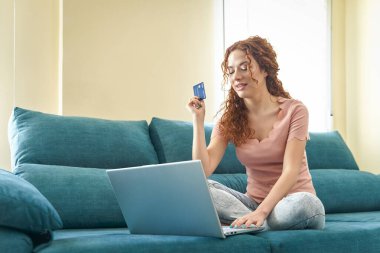 Kafkasyalı, sıradan giyinmiş genç bir kadın oturma odasında oturuyor, kredi kartı ve akıllı telefon kullanarak internetten alışveriş yapıyor. İnternetten alışveriş, online ödeme
