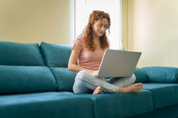 Leende ung kvinna som använder bärbar dator, sitter på soffan hemma, vacker flicka shopping eller chatta online i sociala nätverk, frilansare som arbetar med datorprojekt . — Stockfoto