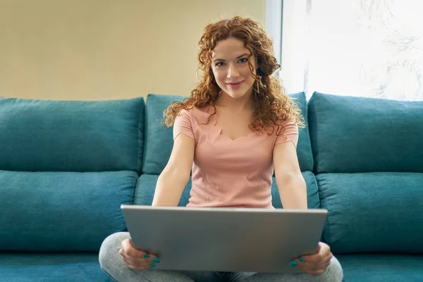 Leende ung kvinna som använder bärbar dator, sitter på soffan hemma, vacker flicka shopping eller chatta online i sociala nätverk, frilansare som arbetar med datorprojekt . — Stockfoto