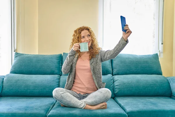 Щаслива руда дівчина бере селфі зі смартфоном і п'є каву. Молода жінка розважається сидячи на дивані вдома . — стокове фото