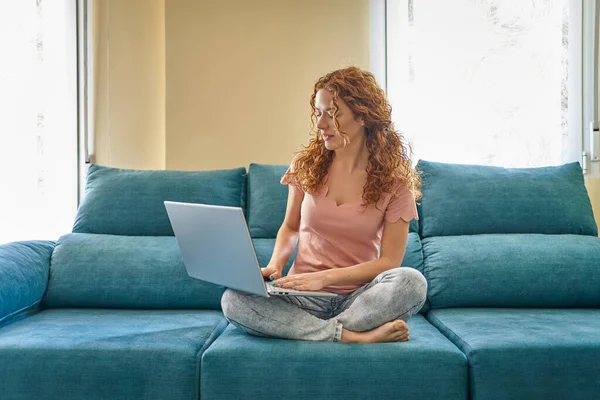 Leende ung kvinna som använder bärbar dator, sitter på soffan hemma, vacker flicka shopping eller chatta online i sociala nätverk, frilansare som arbetar med datorprojekt . — Stockfoto