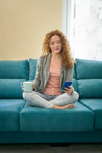 Mujer joven caucásica acostada en un cómodo sofá en una habitación elegante y leyendo noticias con publicación en redes sociales en teléfonos inteligentes modernos utilizando la conexión wifi en el hogar.Hipster blogger descansando en el sofá —  Fotos de Stock