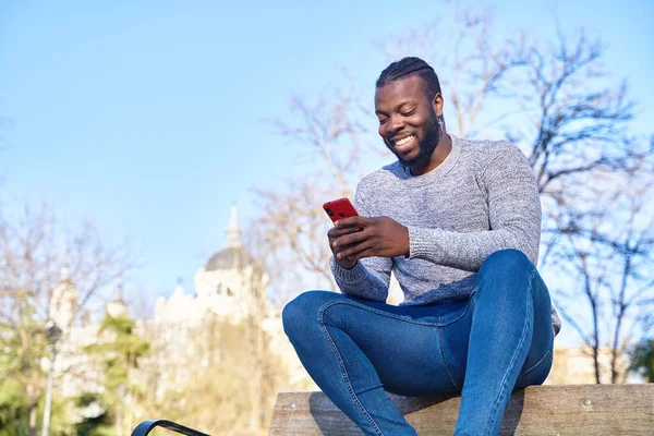 Sorrindo homem negro sentado no banco no parque e usando seu smartphone, espaço de cópia — Fotografia de Stock