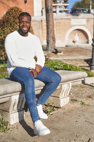 Pria Afrika-Amerika bahagia yang duduk di taman mengenakan jeans, sweater putih dan sepatu putih. Konsep gaya hidup — Stok Foto