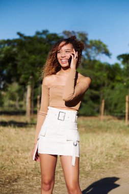 Gülümseyen ve cep telefonu kullanan mutlu Afro-Amerikan kadın. Yeşil arka plan. Bahar ya da yaz mevsimi. Açık havada günlük giysiler.