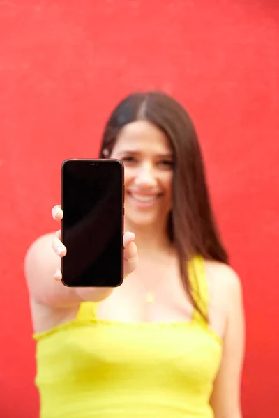 Menina sorrindo mostrando telefone celular com parte superior amarela no fundo vermelho. Foco seletivo no telefone inteligente — Fotografia de Stock