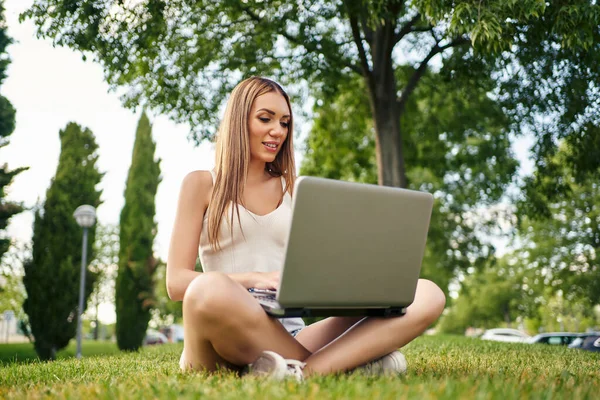 Jovem feliz, sentada na grama, conversando com a família usando laptop, sorrindo amplamente olhando para a câmera contra o fundo do jardim público. Conceito de pessoas e tecnologia. — Fotografia de Stock