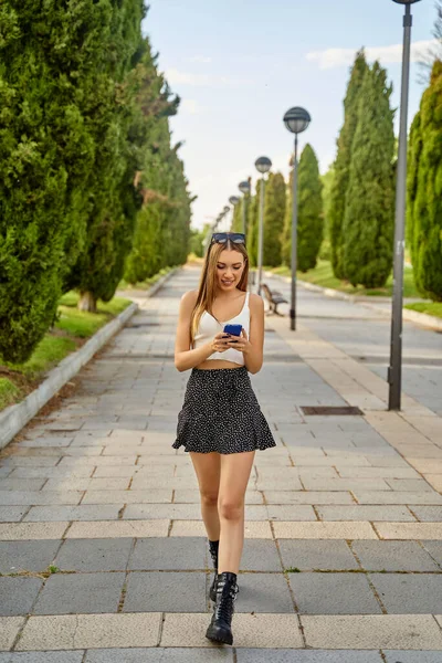 Chica atractiva en ropa de verano caminando en un parque y charlando en su teléfono móvil. Navegar por Internet, ver las redes sociales con su teléfono inteligente. Concepto de tecnología. —  Fotos de Stock