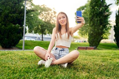 Mutlu gülümseyen sarışın kadın cep telefonuyla, açık havada selfie çekiyor. Tatil yaşam tarzı kavramı. Parkta akıllı telefonu olan güzel bir kız. Güneşli bir gün. Mutluluk kavramı.
