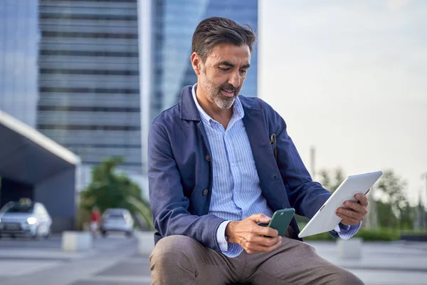 Businessman reading email received on a modern tablet while sitting outdoors in front of an office building, Caucasian man checking a message on a digital touch panel in the city center.