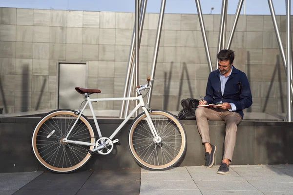 Pengusaha dewasa duduk di buku catatan menulis di buku catatan. Dia menggunakan sepeda untuk berkeliling kota ini. Konsep transportasi hijau. — Stok Foto