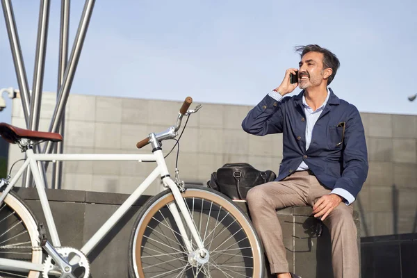 Pria dewasa duduk berbicara di telepon dengan sepeda bersandar di dinding. Konsep transportasi ekologi. — Stok Foto