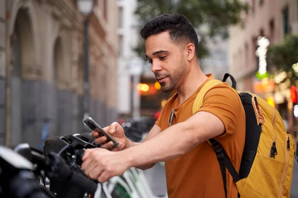 Позитивный молодой латиноамериканец стоит у парковки с велосипедом напрокат и пользуется смартфоном с улыбкой на лице. Экологичная транспортная концепция — стоковое фото