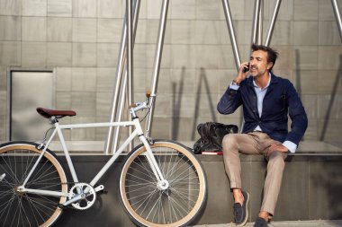 Yetişkin bir erkek telefonda konuşurken duvara yaslanmış bisikletle konuşuyor. Ekolojik ulaşım kavramı. 