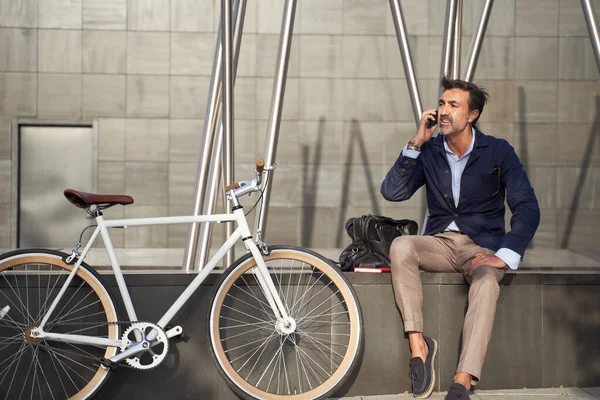 Uomo adulto seduto a parlare al telefono con la bicicletta appoggiata al muro. Concetto di trasporto ecologico. — Foto Stock