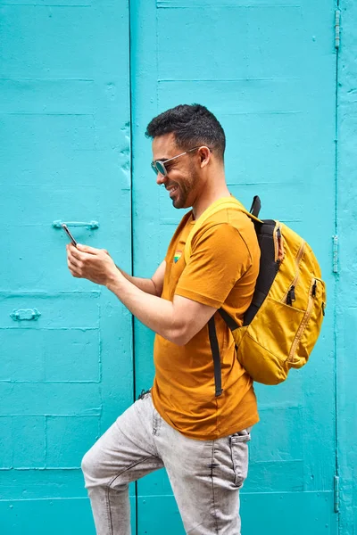 Jovem latino alegre com sua mochila digitando em seu celular andando pela rua, vestindo camiseta laranja e óculos de sol. — Fotografia de Stock