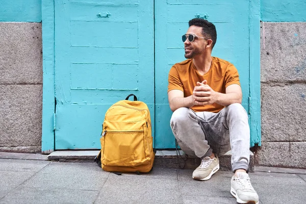 Jovem latino feliz sentado com óculos de sol uma mochila de um lado. Descansando de um passeio na cidade. — Fotografia de Stock