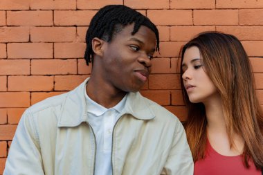 Çok ırklı genç bir çift birbirine bakıyor. Tuğladan bir arka planı var. Sevgi ve bağlılık duygularını ifade ediyorlar.