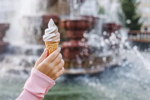 A cone with soft creamy ice cream in children\'s hands on the background of the fountain. In summer, the child walks in the park and eats sweets. Creamy desserts. Horizontal photo.